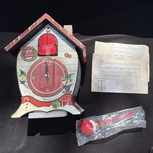 May include: A red and white cuckoo clock shaped like a house with a Santa's Workshop theme. The clock face has a red background with black numbers and hands. There are two elves on either side of the clock face. The clock has a red door that opens and closes. The clock comes with a red and white pendulum.