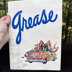 May include: A white rectangular poster featuring the word Grease in large blue cursive script at the top. Below the text is a colorful illustration of a group of people riding in a red convertible car decorated with yellow and orange flame decals.