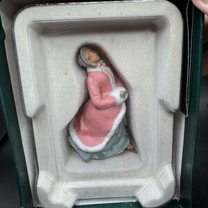Peut inclure: Figurine d'une femme vêtue d'un manteau rose avec une bordure blanche et d'une robe bleue, tenant un petit bouquet. La figurine est à l'intérieur d'un insert en plastique blanc, dans une boîte verte.