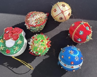 May include: A collection of six vintage Christmas ornaments. The ornaments are various shapes and colors, including red, green, blue, and cream. They are decorated with beads, sequins, and floral designs. Some have gold string hangers.