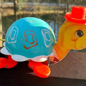 May include: A vintage toy turtle with a blue shell decorated with white and orange designs. The turtle has a yellow head with a red hat and red feet. The toy is made of wood and plastic.