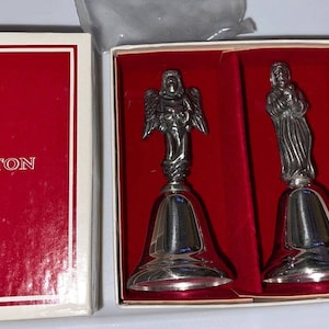 May include: Two silver bells with angel and Mary figures on top. The bells are in a red velvet box with a white outer box. The box has the Reed & Barton logo and the text "The 'Nativity Bells'" on the front. An annual collector's series of tin bells by Reed & Barton.
