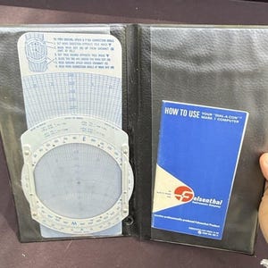 May include: Black leather flight computer case open to reveal a circular slide rule and instruction card. The slide rule has a rotating dial and various scales. A blue booklet with the title "HOW TO USE" is also visible.