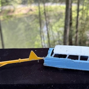 May include: A blue and white toy station wagon with a yellow trailer attached. The toy car and trailer are on a dark surface.