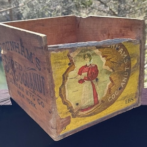 May include: An antique wooden box with the text "DUNHAM'S SHRED COCOANUT" and an illustration of a woman in a red dress. The box is weathered, showing signs of age and use, with peeling labels.