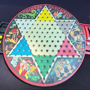 Puede incluir: Un juego de mesa de damas chinas vintage con una superficie de juego en forma de estrella. El tablero presenta secciones triangulares coloridas e ilustraciones. Incluye una bandeja negra y roja con piezas de juego amarillas, azules y blancas.