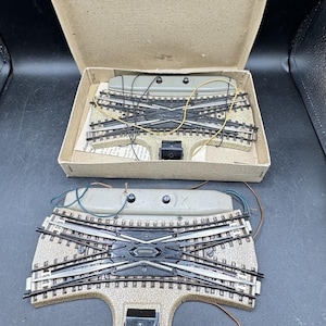 May include: Model train track components, including a crossover and a cardboard box. The tracks are black and silver, with a tan base. Wires and electrical components are visible. The box is open, revealing the tracks.