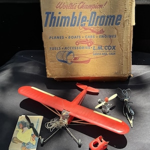 Może przedstawiać: Zestaw modelu samolotu Thimble-Drome w stylu vintage. Zestaw zawiera czerwony model samolotu, silnik, śmigło i akcesoria. Kartonowe pudełko z napisem "World's Champion! Thimble-Drome" i ilustracjami.