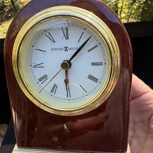 May include: A Howard Miller wooden mantel clock with a gold-tone bezel and Roman numeral hour markers. The clock has a dark brown finish and a gold-tone base. The clock face is white with black hands.