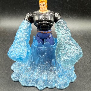 May include: Action figure of a muscular man with orange hair, wearing a black top, blue pants, and a brown belt. The figure has translucent blue water-like arms and is standing in a blue water base. The figure is a collectible toy.
