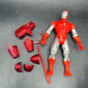 Puede incluir: Una figura de acci&oacute;n de Iron Man roja y plateada con piezas desmontables, incluyendo una pistola, un casco y una armadura para las piernas. La figura est&aacute; en posici&oacute;n vertical, con un dise&ntilde;o detallado y acentos met&aacute;licos. Las piezas est&aacute;n dispersas.