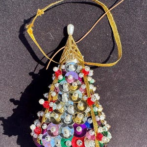 Puede incluir: Adorno de árbol de Navidad hecho a mano. El árbol tiene forma de cono, cubierto de lentejuelas de colores y adornado con pequeñas perlas y cuentas rojas. Tiene una estructura de alambre dorado y una cinta dorada para colgar.