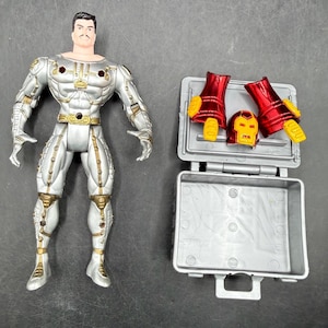 May include: A silver and gold action figure of a superhero with a mustache, standing next to an open gray case containing red and yellow armor pieces and a helmet. The figure has a light skin tone and black hair.