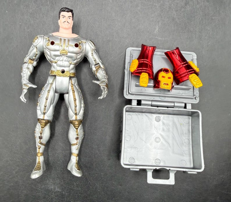 May include: A silver and gold action figure of a superhero with a mustache, standing next to an open gray case containing red and yellow armor pieces and a helmet. The figure has a light skin tone and black hair.