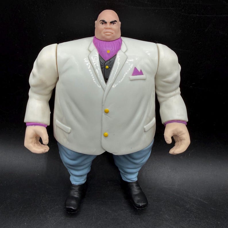 May include: A large action figure of a bald man wearing a white suit jacket with a purple pocket square and trim, a black vest, and light blue pants. The figure has black shoes and a stern expression.
