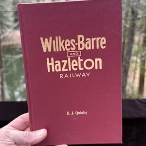 Puede incluir: Un libro granate con el título "Wilkes-Barre and Hazleton Railway" en letras doradas. El nombre del autor, E. J. Quinby, está impreso en la parte inferior. El libro se sostiene en una mano sobre un fondo borroso al aire libre.