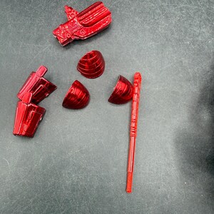 Puede incluir: Un conjunto de piezas de juguete de pl&aacute;stico rojo brillante desmontadas, que incluyen una cabeza, un brazo y secciones esf&eacute;ricas segmentadas, junto con una varilla larga y delgada. Las piezas est&aacute;n dispersas sobre una superficie gris oscuro.