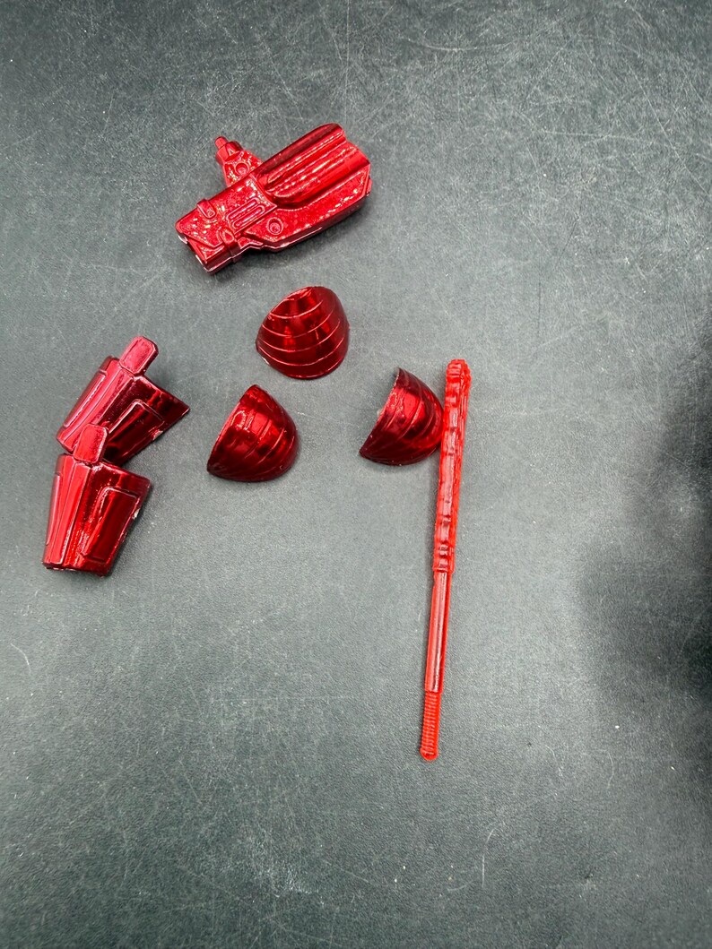Puede incluir: Un conjunto de piezas de juguete de pl&aacute;stico rojo brillante desmontadas, que incluyen una cabeza, un brazo y secciones esf&eacute;ricas segmentadas, junto con una varilla larga y delgada. Las piezas est&aacute;n dispersas sobre una superficie gris oscuro.