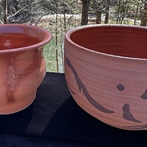 May include: Two handmade terracotta pottery bowls. The bowl on the left is a small, rounded planter with a flared rim. The bowl on the right is a larger, open bowl with a decorative dark brown design. Both bowls have a rustic, earthy aesthetic.