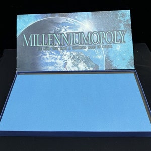 May include: A blue box with a blue felt lining. The lid of the box has a picture of the Earth with the text "Millenniumopoly The Game That Took A Thousand Years To Create."