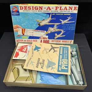 Op de afbeelding: Vintage "Design-A-Plane" modelvliegtuig constructieset. De doos toont illustraties van straaljagers en de tekst "Design-A-Plane" en "Jet Fighter Series". De doos bevat modelonderdelen en instructies.