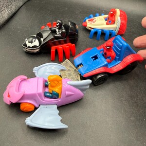 May include: Four toy vehicles featuring superhero characters. Includes a black and red motorcycle with a Venom figure, a red and blue car with a Spider-Man figure, a white and red vehicle with a Spider-Man figure, and a pink aircraft with a figure.