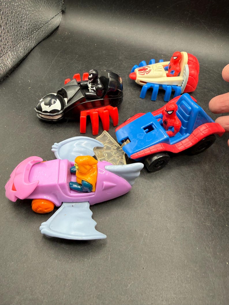 May include: Four toy vehicles featuring superhero characters. Includes a black and red motorcycle with a Venom figure, a red and blue car with a Spider-Man figure, a white and red vehicle with a Spider-Man figure, and a pink aircraft with a figure.