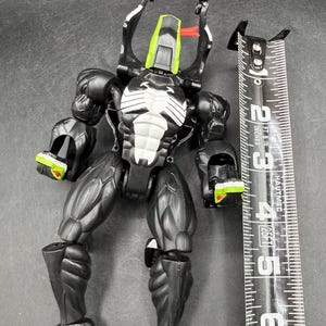May include: A black and white action figure with green and red accents, next to a measuring tape. The figure has a muscular build and detailed design. The measuring tape shows measurements in inches, equivalent to cm.