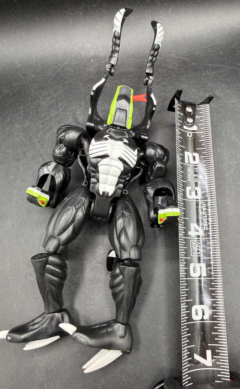 May include: A black and white action figure with green and red accents, next to a measuring tape. The figure has a muscular build and detailed design. The measuring tape shows measurements in inches, equivalent to cm.