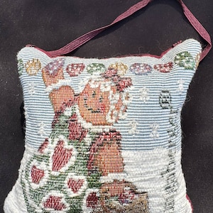 May include: A small, square, white pillow with a red, green, and brown embroidered design of a snowman holding a Christmas tree. The pillow has a burgundy fabric strap attached to the top.