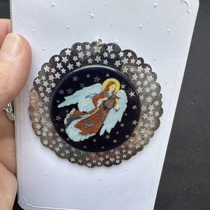 Peut inclure: Ornement rond représentant un ange, avec une bordure argentée en forme d'étoiles. L'ange, aux ailes blanches et à la robe rouge et dorée, est posé sur un fond bleu foncé. Il est fixé à une carte blanche ornée d'étoiles en relief.