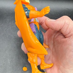 May include: Orange action figure with a cape, boots, and gloves. The figure has a yellow face and is wearing a blue and purple outfit. A small orange piece is detached from the figure.