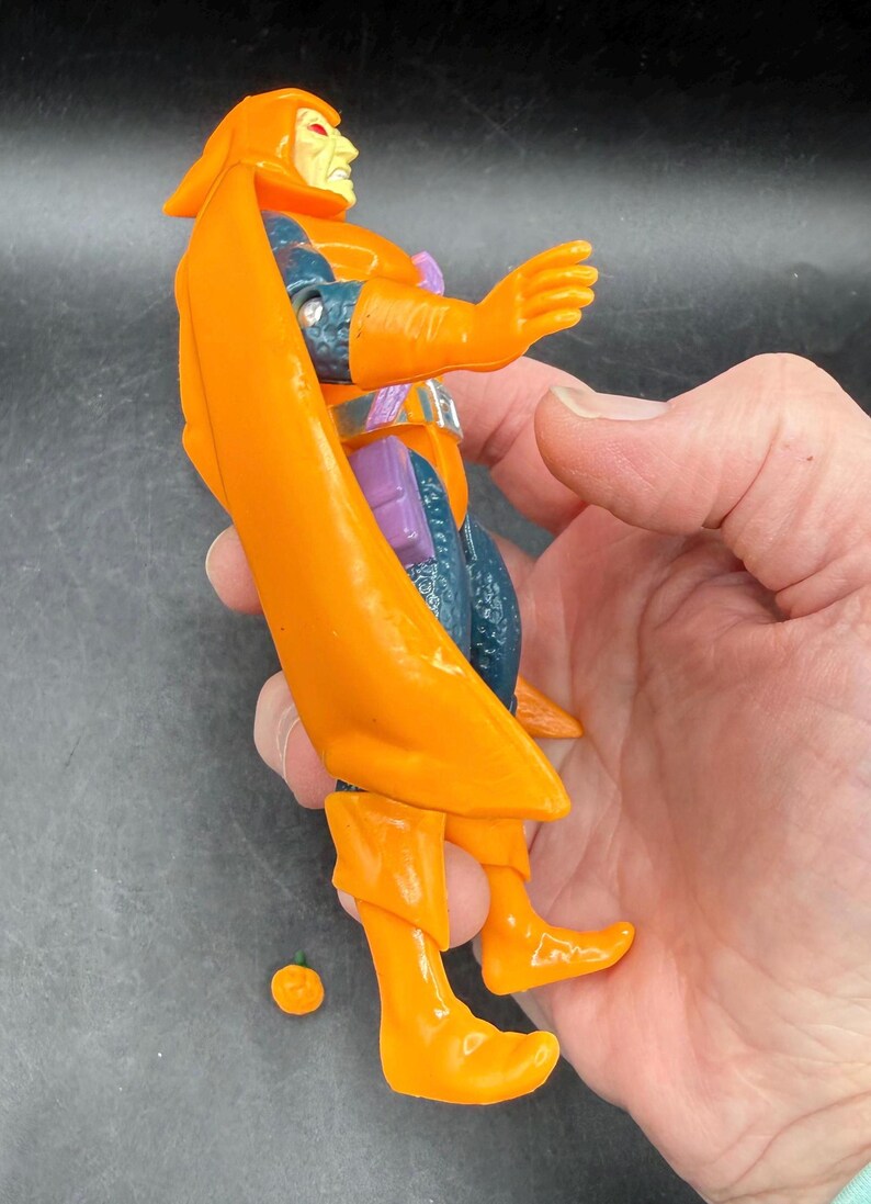 May include: Orange action figure with a cape, boots, and gloves. The figure has a yellow face and is wearing a blue and purple outfit. A small orange piece is detached from the figure.