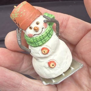 May include: A whimsical snowman figurine with a carrot nose, button eyes, and a smiling mouth. The snowman wears a green scarf, an orange pot-shaped hat, and has twig arms. It is mounted on a small, rectangular base.
