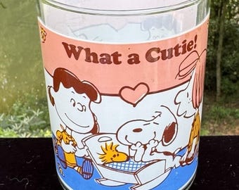 Può includere: Bicchiere di vetro trasparente con un design a cartoni animati dei Peanuts. Il bicchiere ha una fascia rosa con le parole "What a Cutie!" e illustrazioni di Snoopy, Woodstock e altri personaggi. La base è trasparente e il bordo è liscio.