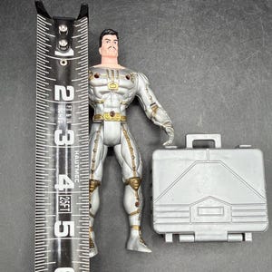 May include: A silver action figure with gold accents, standing next to a silver briefcase. The figure is approximately 5 inches tall, with a detailed design. The figure has a mustache and black hair. The briefcase has a handle and a rectangular shape.