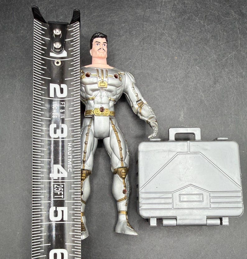 May include: A silver action figure with gold accents, standing next to a silver briefcase. The figure is approximately 5 inches tall, with a detailed design. The figure has a mustache and black hair. The briefcase has a handle and a rectangular shape.