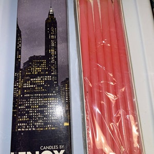 May include: A box of pink taper candles by Lenox. The box features a nighttime cityscape design. The candles are packaged in a clear plastic wrap inside a cardboard box. The box reads "CANDLES BY LENOX WITH PATENTED STANDTITE BASE."