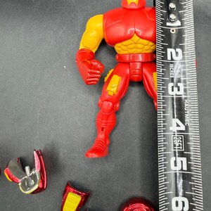 May include: A red and yellow Iron Man action figure, approximately 5 inches tall, with detachable accessories. The figure has a red helmet, chest, and legs, with yellow arms and chest details. Additional red and silver pieces are scattered around.
