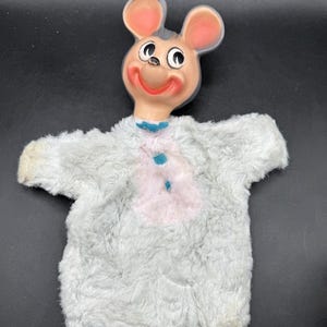 Könnte beinhalten: Vintage Handpuppe einer Maus mit bemaltem Gesicht und großen rosa Ohren. Der Körper der Puppe besteht aus weichem, grauem Fell mit einer rosa Brust und blauen Akzenten. Die Puppe hat einen lächelnden Gesichtsausdruck.