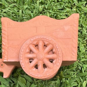 Könnte beinhalten: Ein dekorativer Wagen-Pflanztopf in Terrakotta-Farbe. Der Pflanztopf hat ein detailliertes Wagenrad und eine geschwungene Oberseite. Der Wagen besteht aus einem rötlich-braunen Tonmaterial und ist für Pflanzen oder Blumen konzipiert. Der Pflanztopf steht auf grünem Gras.