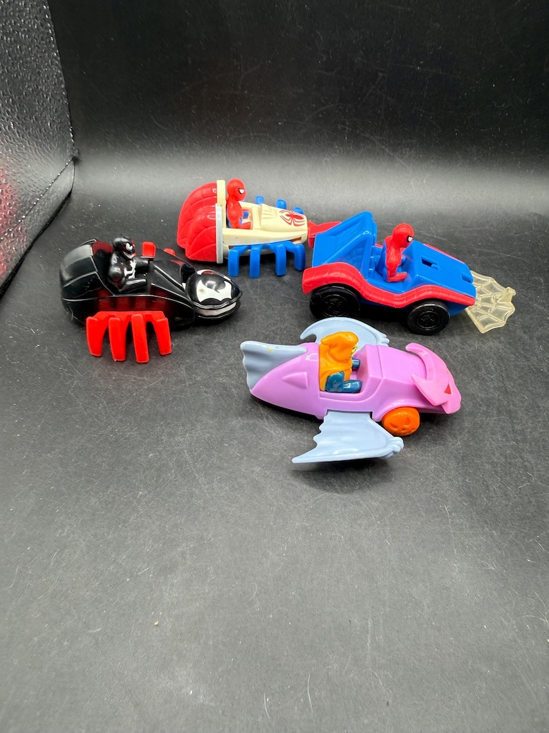 May include: Four toy vehicles featuring Spider-Man characters. Includes a black motorcycle, a red and blue car, a blue and red car with a web, and a pink and blue jet. Each vehicle has a character figure.