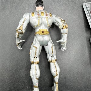 May include: A silver action figure with gold accents, viewed from the back. The figure has a muscular build and is wearing a detailed suit with various panels and joints. The figure's hair is black.