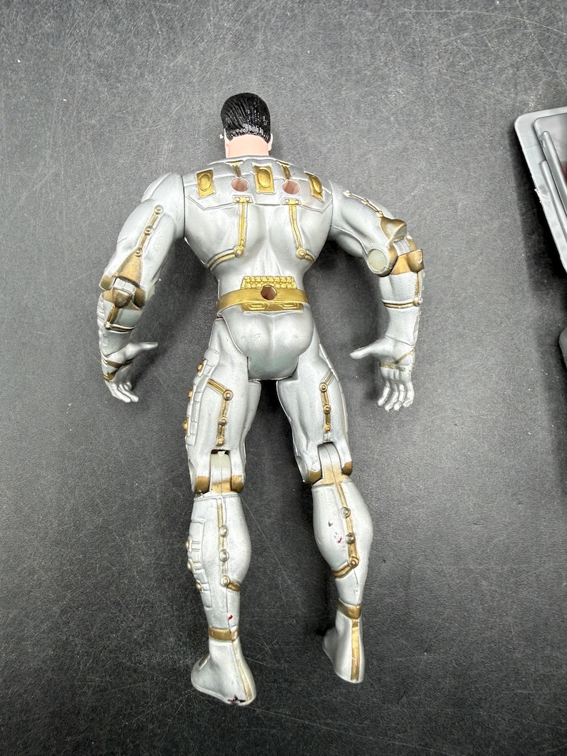 May include: A silver action figure with gold accents, viewed from the back. The figure has a muscular build and is wearing a detailed suit with various panels and joints. The figure's hair is black.