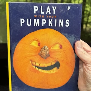 Puede incluir: Un libro titulado "Play With Your Pumpkins" presenta una cara de calabaza sonriente en la portada. La calabaza es naranja con ojos y boca tallados, sobre un fondo azul oscuro. El libro tiene un lomo amarillo.