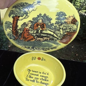 May include: A yellow ceramic trinket box with a lid featuring a pastoral scene with two figures, a tree, and a small house. The inside of the box is yellow and has a quote: "My heart is fix'd I cannot range, I like my choice Too well to change."