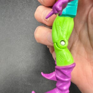 May include: A close-up of a Green Goblin action figure. The figure has a green body, purple boots and gloves, and a teal torso. The figure is held in a hand against a dark background.