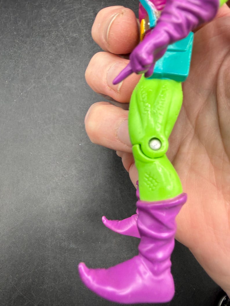 May include: A close-up of a Green Goblin action figure. The figure has a green body, purple boots and gloves, and a teal torso. The figure is held in a hand against a dark background.
