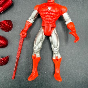Puede incluir: Una figura de acci&oacute;n de Iron Man roja y plateada con un bast&oacute;n rojo. La figura tiene un casco, pecho y botas rojos, con brazos y piernas plateados. Varios accesorios rojos est&aacute;n esparcidos alrededor de la figura.