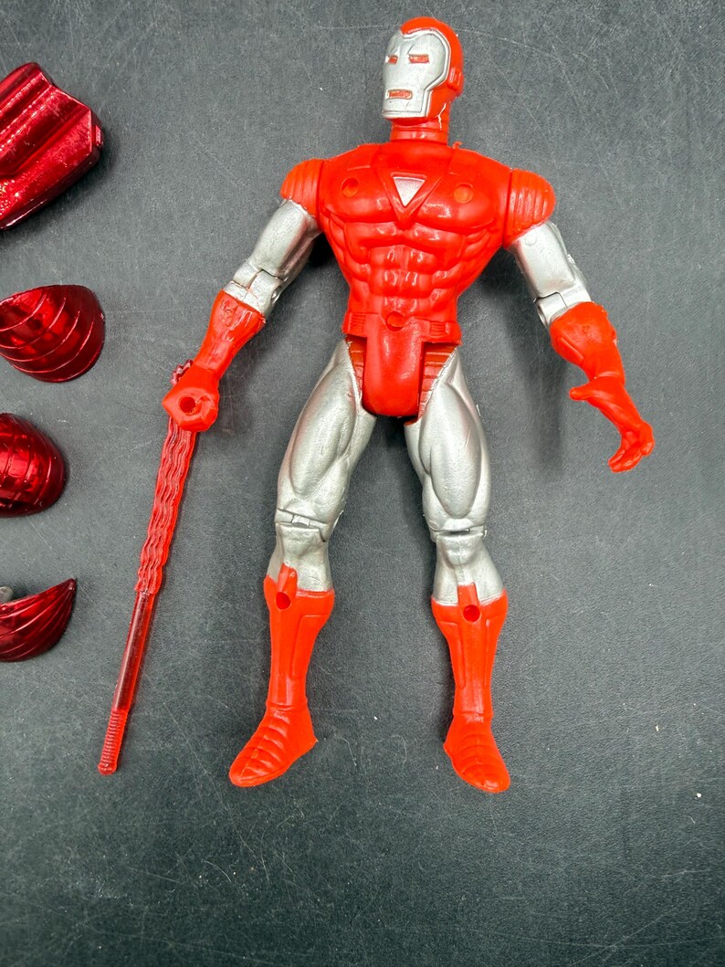 Puede incluir: Una figura de acci&oacute;n de Iron Man roja y plateada con un bast&oacute;n rojo. La figura tiene un casco, pecho y botas rojos, con brazos y piernas plateados. Varios accesorios rojos est&aacute;n esparcidos alrededor de la figura.